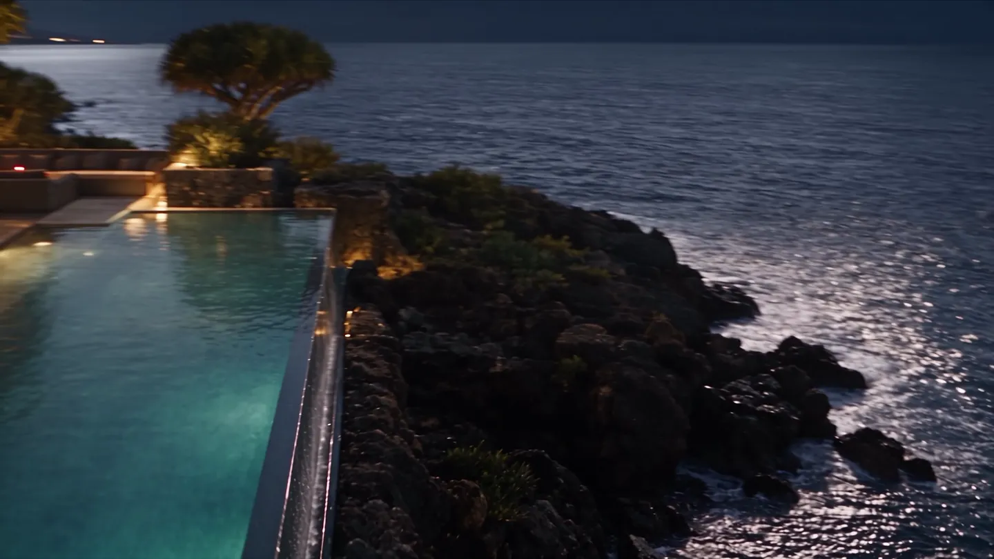 At night, the lit infinity pool on the edge of the cliff and the sea under the moon.